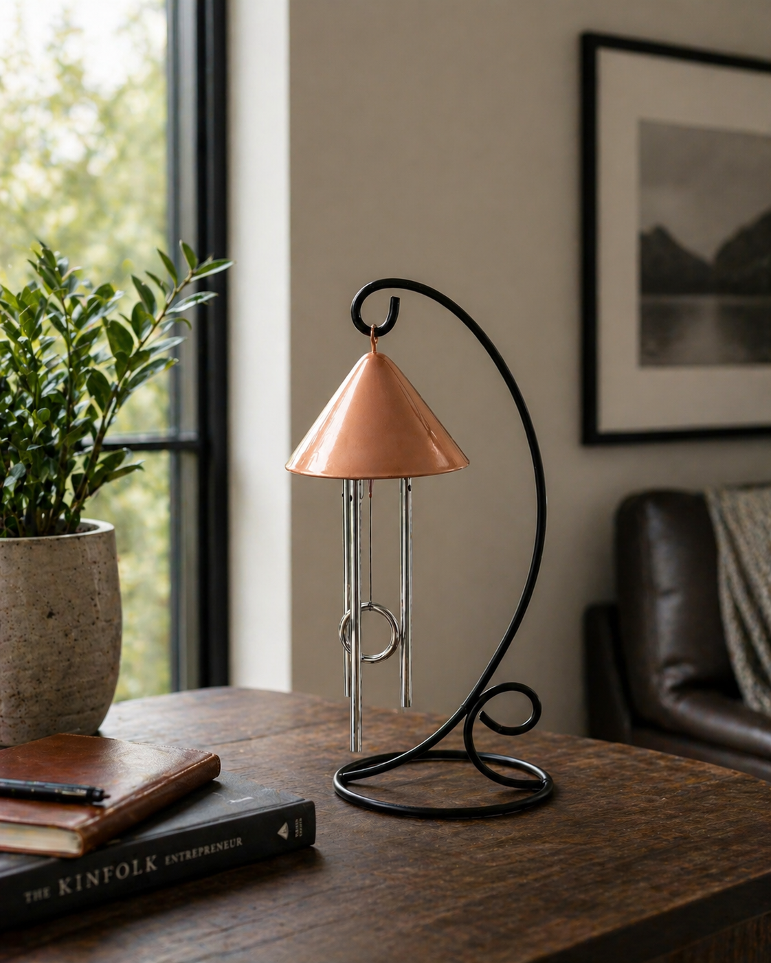 Decorative copper solar wind chime on a wooden table with a plant and books near window.