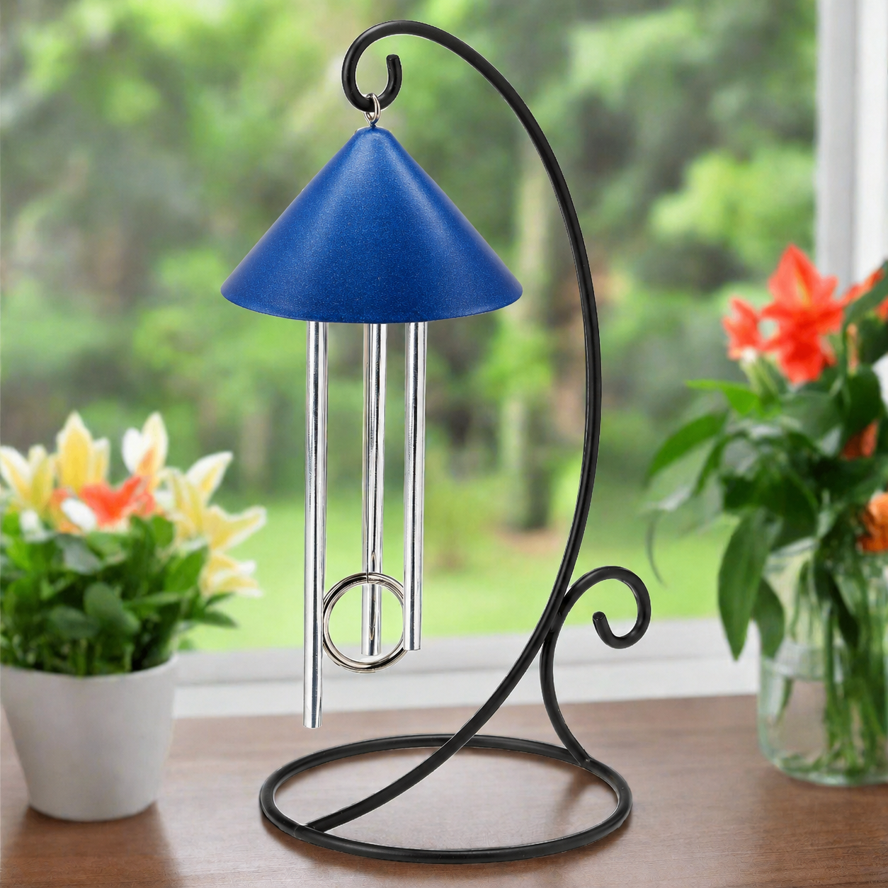Indoor solar wind chime with the royal  blue shimmer shade and silver hanging chime rods, mounted on a black curved scroll-style stand, placed on a wooden table with potted flowers in front of a sunny cheerful window.